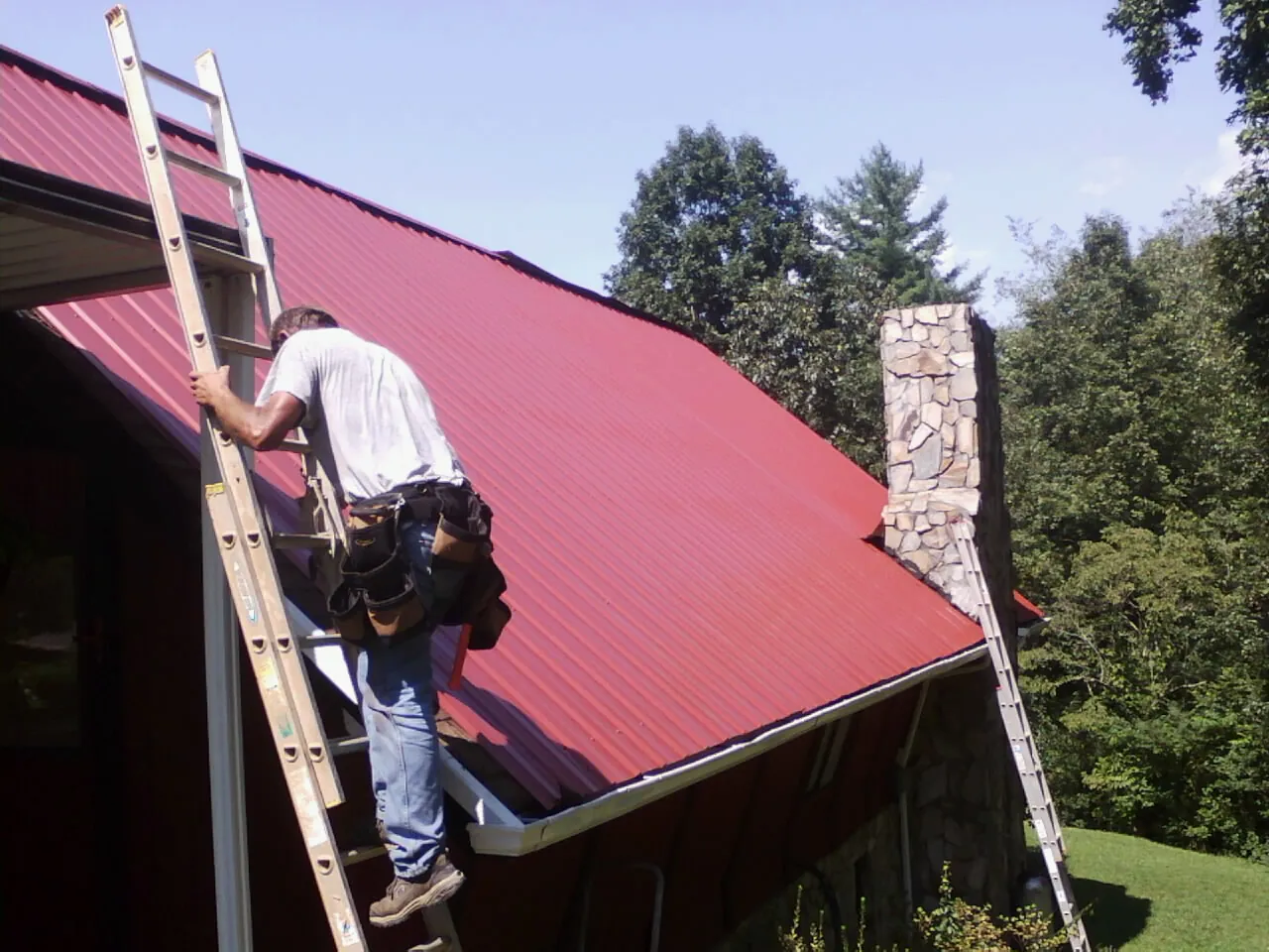 Roofer on a ladder beside a red metal roof and stone chimney — Soffit & Fascia Repair by Roofing Brothers in Chickasha